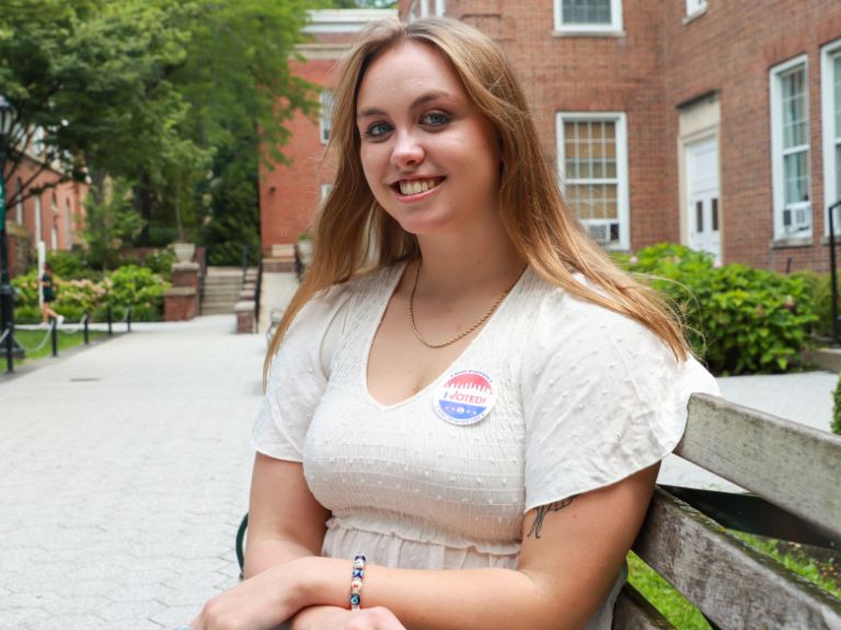 female student with blond hair sitting outside the library with an I Voted sticker